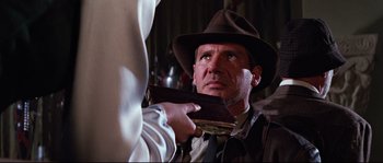 Movie still from “Indiana Jones and the Last Crusade” (1989), directed by Steven Spielberg – A man in a hat holding a wallet and looking at another man in a suit; Close Up shot, Over the shoulder angle