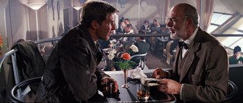 Movie still from “Indiana Jones and the Last Crusade” (1989), directed by Steven Spielberg – Two men sitting at a table in front of each other at a restaurant; Medium shot, Over the shoulder angle