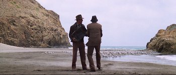 Movie still from “Indiana Jones and the Last Crusade” (1989), directed by Steven Spielberg – A couple of men standing on top of a sandy beach; Wide shot, Low angle