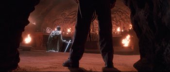 Movie still from “Indiana Jones and the Last Crusade” (1989), directed by Steven Spielberg – A man standing in front of an audience in a dark room; Wide shot, Low angle
