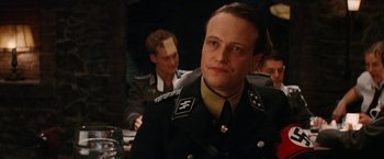 Movie still from “Inglourious Basterds” (2009), directed by Quentin Tarantino – A man in a military uniform is looking at the camera while another man is sitting in the background; Close Up shot, Over the shoulder angle