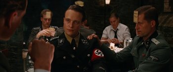 Movie still from “Inglourious Basterds” (2009), directed by Quentin Tarantino – A man in a military uniform sitting in front of a group of people; Close Up shot, Over the shoulder angle