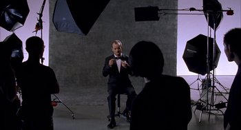 Movie still from “Lost in Translation” (2003), directed by Sofia Coppola – A man in a suit and bow tie sitting in front of a camera; Medium shot, Over the shoulder angle