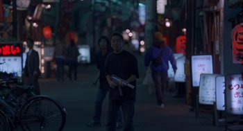 Movie still from “Lost in Translation” (2003), directed by Sofia Coppola – A man standing on a street holding a guitar; Wide shot, High angle