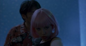 Movie still from “Lost in Translation” (2003), directed by Sofia Coppola – A woman with pink hair is holding a microphone; Medium shot, Over the shoulder angle