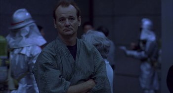 Movie still from “Lost in Translation” (2003), directed by Sofia Coppola – A man with his arms crossed standing in front of a group of people; Medium shot, Over the shoulder angle