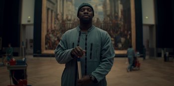 Movie still from “Lupin” (2021), created by Tigran Rosine – A man standing in front of a painting holding a book; Medium shot, Low angle
