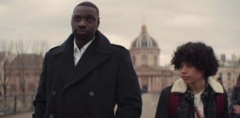 Movie still from “Lupin” (2021), created by Tigran Rosine – A man and a woman standing next to each other in front of a building; Medium shot, Over the shoulder angle