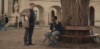 Movie still from “Lupin” (2021), created by Tigran Rosine – Two men standing next to each other near a tree; Medium shot, Over the shoulder angle