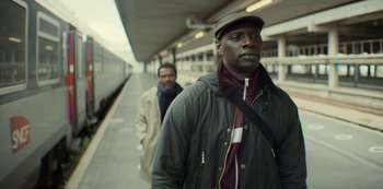 Movie still from “Lupin” (2021), created by Tigran Rosine – Two men are walking on a train platform; Medium shot, Over the shoulder angle