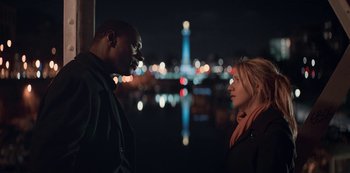 Movie still from “Lupin” (2021), created by Tigran Rosine – A man and a woman standing next to each other in front of a city skyline at night; Medium shot, Low angle