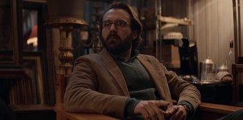 Movie still from “Lupin” (2021), created by Tigran Rosine – A man sitting in a chair wearing glasses and a brown jacket; Medium shot, Low angle