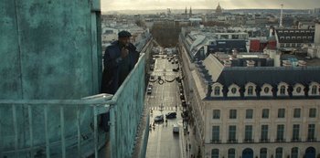 Movie still from “Lupin” (2021), created by Tigran Rosine – A man standing on the edge of a building looking down at a street; Extreme Wide shot, Low angle