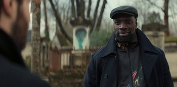 Movie still from “Lupin” (2021), created by Tigran Rosine – A man wearing a hat standing in front of a statue; Medium shot, Over the shoulder angle
