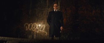 Movie still from “Kingsman: The Secret Service” (2014), directed by Matthew Vaughn – A man in a black coat and tie standing in front of a brick wall; Wide shot, Low angle
