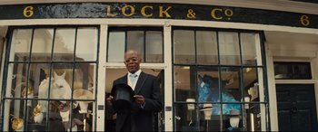 Movie still from “Kingsman: The Secret Service” (2014), directed by Matthew Vaughn – A man standing in front of a store holding a hat; Medium shot, Low angle