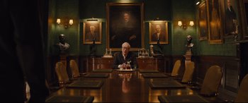 Movie still from “Kingsman: The Secret Service” (2014), directed by Matthew Vaughn – A man sitting at a table in front of a painting; Wide shot, Low angle