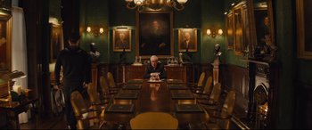 Movie still from “Kingsman: The Secret Service” (2014), directed by Matthew Vaughn – An older man sitting at a large wooden table in front of a painting; Wide shot, Over the shoulder angle