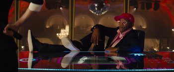 Movie still from “Kingsman: The Secret Service” (2014), directed by Matthew Vaughn – A man sitting on top of a glass table in a room; Medium shot, Low angle
