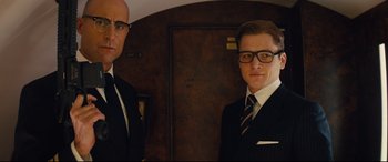 Movie still from “Kingsman: The Secret Service” (2014), directed by Matthew Vaughn – A man in a suit standing next to another man in a suit; Medium shot, Low angle