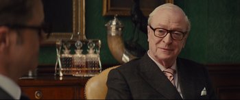 Movie still from “Kingsman: The Secret Service” (2014), directed by Matthew Vaughn – An older man sitting in a chair in front of a table; Close Up shot, Over the shoulder angle