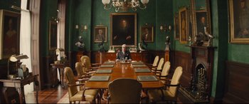 Movie still from “Kingsman: The Secret Service” (2014), directed by Matthew Vaughn – A man sitting at a large wooden table in front of a painting; Wide shot, Low angle