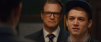 Movie still from “Kingsman: The Secret Service” (2014), directed by Matthew Vaughn – A man in a suit standing next to another man in a baseball cap; Close Up shot, Over the shoulder angle