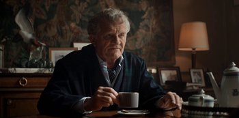 Movie still from “Lupin” (2021), created by Tigran Rosine – An older man sitting at a table with a cup of coffee; Medium shot, Low angle