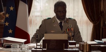 Movie still from “Lupin” (2021), created by Tigran Rosine – A man sitting at a table wearing a military uniform; Medium shot, Low angle