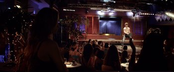 Movie still from “Magic Mike” (2012), directed by Steven Soderbergh – A group of people sitting at tables in front of a microphone; Extreme Wide shot, Over the shoulder angle