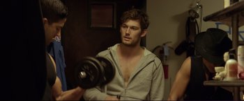 Movie still from “Magic Mike” (2012), directed by Steven Soderbergh – A young man in a gray hoodie is holding a black dumbbell; Medium shot, Over the shoulder angle