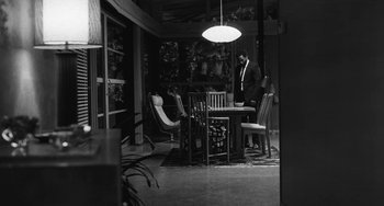Movie still from “Malcolm & Marie” (2021), directed by Sam Levinson – A black and white photo of a man sitting at a dining room table; Wide shot, High angle