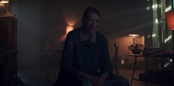 Movie still from “Marianne” (2019), created by Samuel Bodin – An older woman sitting on a chair in a room; Medium shot, Over the shoulder angle