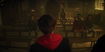 Movie still from “Marianne” (2019), created by Samuel Bodin – A group of people sitting on benches in a dark room; Wide shot, Over the shoulder angle