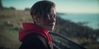 Movie still from “Marianne” (2019), created by Samuel Bodin – A young man with a red hooded sweatshirt looks into the distance while standing in front of the ocean; Close Up shot, Over the shoulder angle