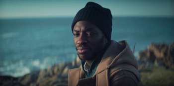 Movie still from “Marianne” (2019), created by Samuel Bodin – A man wearing a coat and a hat near the ocean; Close Up shot, High angle