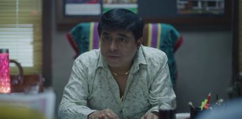 Movie still from “Maska” (2020), directed by Neeraj Udhwani – A man sitting at a table in front of a colorful chair; Close Up shot, Low angle