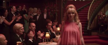 Movie still from “Last Night in Soho” (2021), directed by Edgar Wright – A group of people sitting at a table with a woman in a pink dress in front of them; Medium shot, Over the shoulder angle