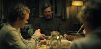 Movie still from “Midnight Mass” (2021), directed by Mike Flanagan – A man sitting at a table with a plate in front of him; Medium shot, Over the shoulder angle