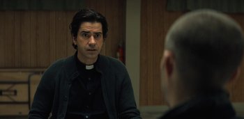 Movie still from “Midnight Mass” (2021), directed by Mike Flanagan – A man wearing a priest's collar is talking to another man in front of a wall; Close Up shot, Over the shoulder angle