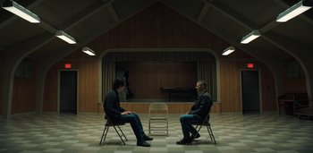 Movie still from “Midnight Mass” (2021), directed by Mike Flanagan – Two men sitting in chairs facing each other in front of a white chair; Wide shot, High angle
