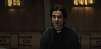 Movie still from “Midnight Mass” (2021), directed by Mike Flanagan – A man in a priest's outfit sitting in a room; Close Up shot, Over the shoulder angle