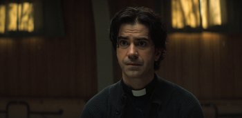 Movie still from “Midnight Mass” (2021), directed by Mike Flanagan – A man in a priest's outfit stares into the camera; Close Up shot, Over the shoulder angle