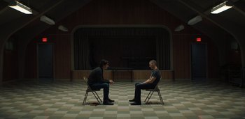 Movie still from “Midnight Mass” (2021), directed by Mike Flanagan – Two men sitting in chairs facing each other in front of a stage; Wide shot, Low angle