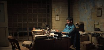 Movie still from “Midnight Mass” (2021), directed by Mike Flanagan – A man sitting at a desk in front of a laptop computer; Medium shot, High angle