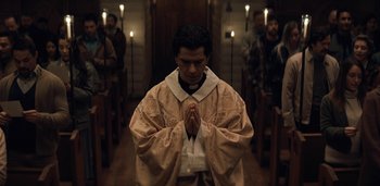 Movie still from “Midnight Mass” (2021), directed by Mike Flanagan – A man in a priest's outfit praying in front of a group of people; Medium shot, Low angle