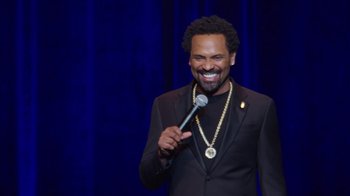 Movie still from “Mike Epps: Only One Mike” (2019), directed by Kevin Bray – A man in a black suit holding a microphone; Medium shot, Over the shoulder angle