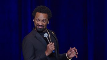 Movie still from “Mike Epps: Only One Mike” (2019), directed by Kevin Bray – A man holding a microphone in front of a blue curtain; Close Up shot, Over the shoulder angle