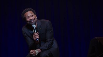 Movie still from “Mike Epps: Only One Mike” (2019), directed by Kevin Bray – A man sitting on a stage holding a microphone; Medium shot, High angle