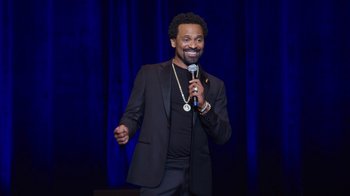 Movie still from “Mike Epps: Only One Mike” (2019), directed by Kevin Bray – A man holding a microphone while wearing a suit; Medium shot, Overhead angle
